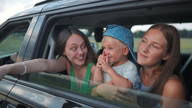 Two women smile with playful boy at car window during road trip. Happy family enjoys countryside journey. Child plays between women. Summer vacation in car. Boy and women share fun trip.