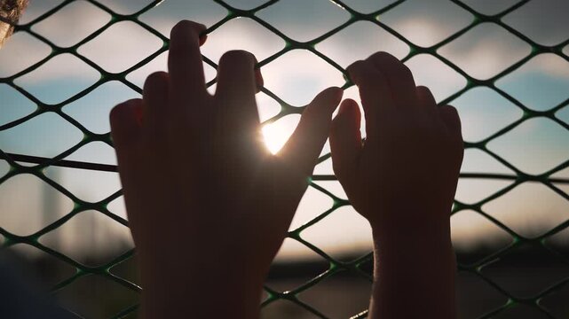 Hands gripping chainlink fence at sunset with fingers through mesh. Person holds fence barrier outdoors. Silhouette hands on chainlink at sunset. Fingers grip wire fence with sunlight behind.