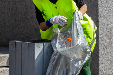 Worker in a bright neon-yellow safety vest and protective gloves is carefully emptying a transparent garbage bag filled with waste into a metal public bin. The scene highlights urban cleaning efforts,