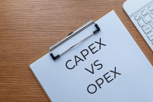 A clipboard on a wooden desk displaying CAPEX VS OPEX text. Business finance and accounting concepts regarding capital and operating expenditures.