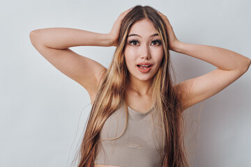 Fototapeta premium Young woman with long hair and surprised expression holding head with hands on white background, casual sleeveless top and natural makeup closeup portrait.