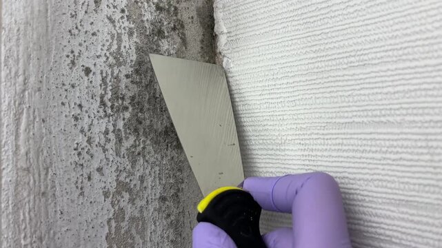 A man wearing gloves removes mold from a wall using a putty knife. 