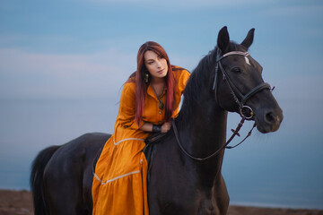 Obraz premium A woman with beautiful hair in an orange dress tenderly hugs a horse against the backdrop of a calm sky and a lake. Photo shoot by the water.