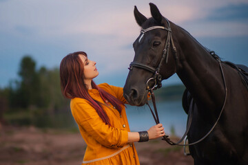 Obraz premium A woman with beautiful hair in an orange dress tenderly hugs a horse against the backdrop of a calm sky and a lake. Photo shoot by the water.