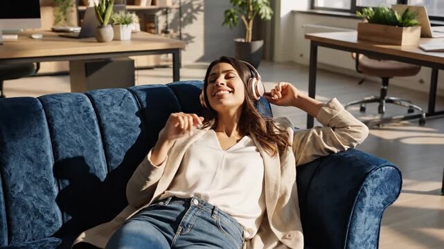 A female entrepreneur listens to music while lying down on a blue sofa in her office. She smiles and relaxes in a well-lit space with plants and workstations nearby