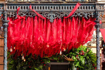 Rich red fabric cascades from an intricately designed wrought iron framework, enhanced by warm hanging light bulbs and lush greenery below. The scene blends vintage charm with festive flair, creating 
