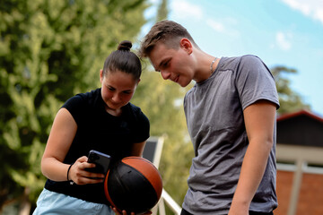 Fototapeta premium Teenage basketball players engage in friendly conversation while reviewing sports clips on a mobile device at an outdoor court during a sunny afternoon in a park
