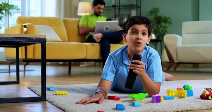 Indian asian boy watching TV lying on carpet while father working on laptop on sofa behind him in modern home, showing everyday family lifestyle
