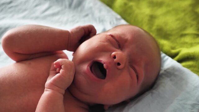 A close-up portrait of a week-old infant crying. The baby's face is scrunched up, mouth wide open, resting on soft blue and green blankets. Authentic, candid lifestyle moment