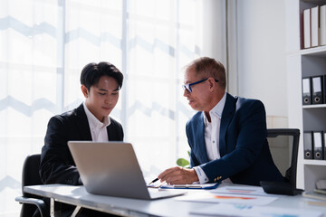 Fototapeta premium Two businessmen are discussing a project, with one older male pointing to a laptop screen while a younger male listens intently, showing teamwork and mentorship in an office setting