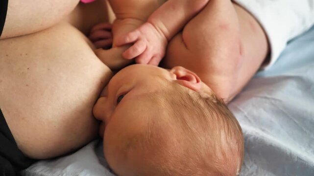High-angle close-up of a newborn baby nursing. The infant's head, fine hair, and tiny ear are visible while nestled against mother's breast. Soft, natural light creates a tender mood