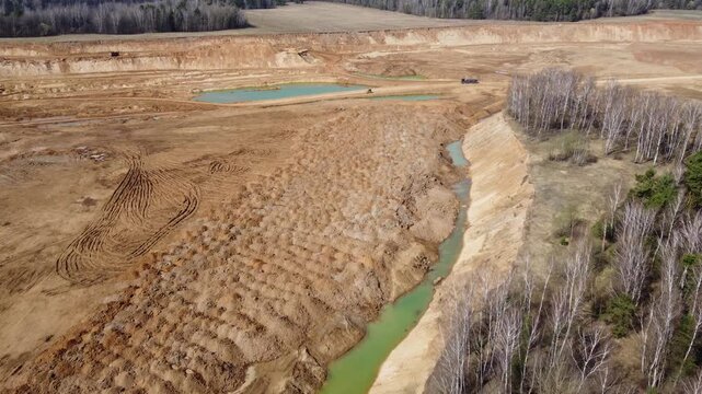 Aerial view captures vast brown sand quarry excavation adjacent to a thin ribbon of turquoise water and encroaching white birch forest