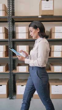 Woman entrepreneur reviewing inventory on a tablet while walking through a storage room filled with organized boxes, vertical shot