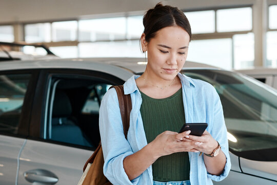 Asian woman, typing and phone at car dealership to sale, customer and website for research. Female person, communication and mobile app for vehicle review or online survey, showroom and social media