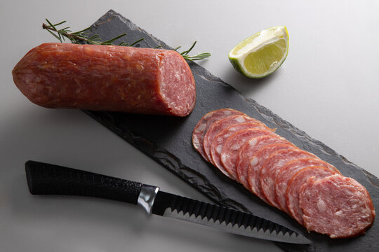 Salame fatiado e inteiro apresentado sobre uma placa de ard&oacute;sia escura, com uma faca serrilhada, um ramo de alecrim e uma fatia de lim&atilde;o. Composi&ccedil;&atilde;o minimalista de comida em est&uacute;dio