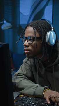 Vertical of Black male cybercriminal wearing headphones and glasses typing on computer focused on coding breach into secured servers during evening in hidden home office