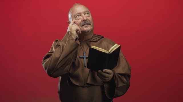 Man monk with finger to temple holding open bible and wooden cross necklace in studio; calm contemplation.
