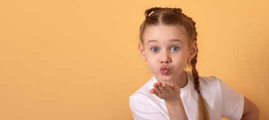 Adorable young girl in white t-shirt blowing a kiss with playful expression, isolated on pastel orange background, copy space.