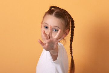 Portrait of a young girl with braids playfully extending her hand towards the camera against a warm orange background.