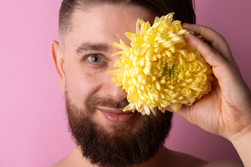 Smiling bearded man covering one eye with a bright yellow chrysanthemum against a pastel pink background.