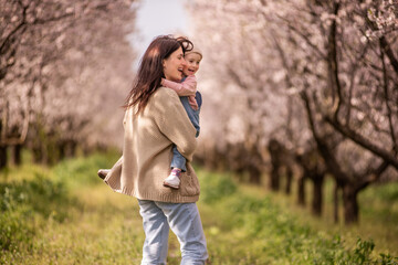 Mother carries smiling daughter through blooming almond orchard, sharing a joyful spring moment of bonding and warmth among green grass and sunlit trees