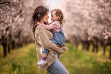 Mother holding her young daughter close, showing a bond of love and family connection in a beautiful spring orchard with trees full of delicate white blossoms