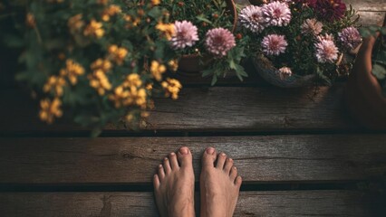 Fototapeta premium Bare feet stand on a weathered wooden deck beside baskets of colorful flowers. Concept Bare feet on weathered deck, Colorful flower baskets, Rustic outdoor setting, Sunlit natural textures