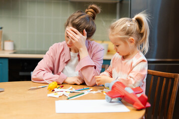Mother experiencing stress and headache while daughter plays with toys in kitchen