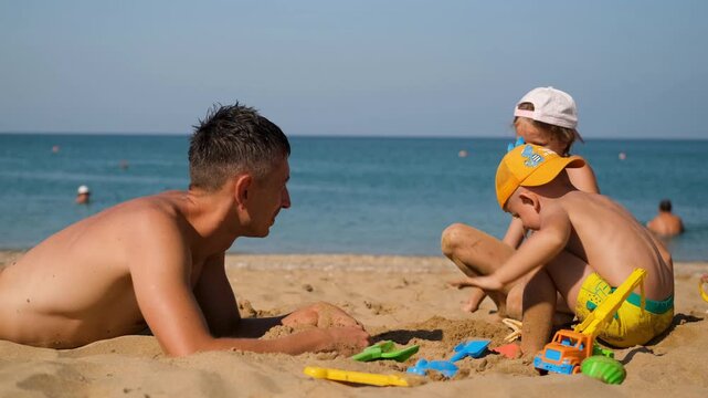A little boy slaps his hands in the sand on the beach. His father and sister look on with smiles.