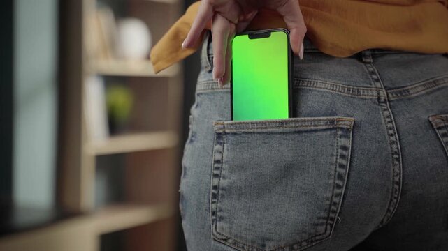 Person inserting a mobile device with a customizable green screen display into the back pocket of blue denim jeans, symbolizing modern technology, convenience, and everyday digital integration