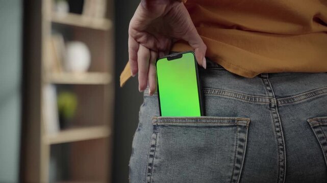 Woman's hand placing a mobile phone with a customizable green chroma key screen into the back pocket of her blue denim jeans, highlighting privacy and device accessibility