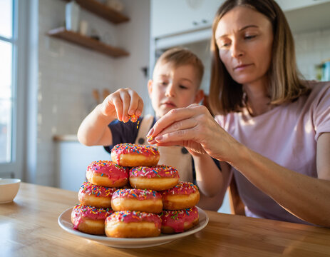Mama i syn dekorujący stos pączk&oacute;w leżących na talerzu kolorowymi posypkami
