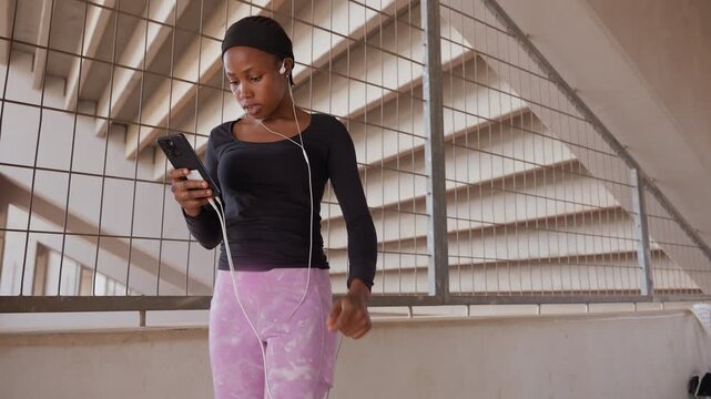 black woman checking phone on balcony, standing on concrete landing under architectural beams, scrolling messages and glancing outward, purple pants and black top, quiet contemplative mood,