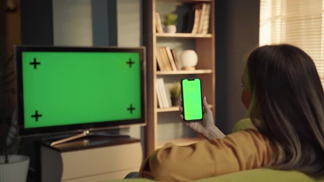 Woman on sofa in living room holding smartphone with blank chroma key screen while tv also shows green screen, illustrating multi device connectivity and media consumption at home