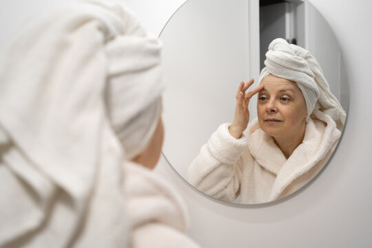 Middle-aged woman looks at herself in bathroom mirror with towel on head.