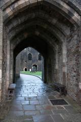 Fototapeta premium A wide-angle interior view looking through a massive stone vaulted archway of a medieval castle, leading to a cobblestone courtyard with wooden benches and historic ruins in the background.