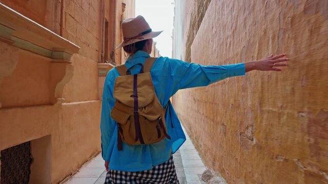 A female tourist in a hat and with a backpack walks along the traditional narrow old streets in Mdina, Malta