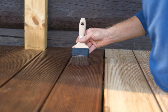 Brown oil for protection of wood by staining. Painting a larch for protecting and pests. Woodworker applying finish to terrace deck board. Repairman varnishing with brush wooden plank floor surface.