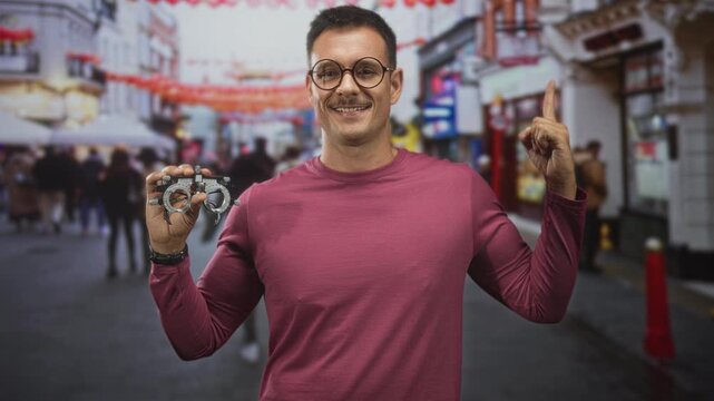Man holding a trial frame with his hand and points finger upward while smiling in a busy street lined with shops and lanterns; confidence.