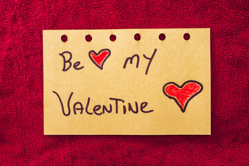 Be My Valentine, written on a piece of paper, on a red textile background.
