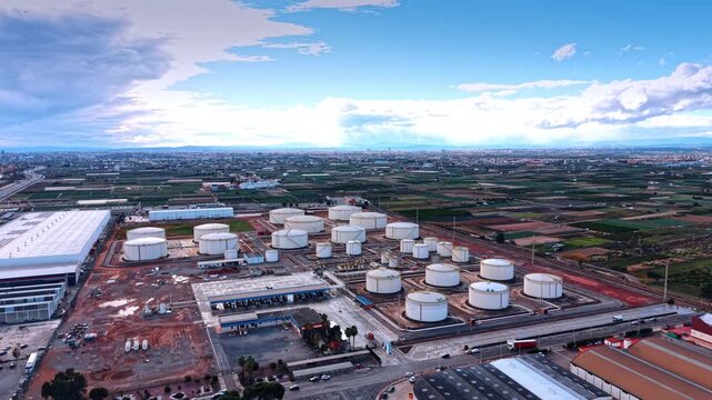 Approaching a large territory of a modern oil refinery plant with diverse size tanks for fuel storage. Aerial view. Green fields and vast cityscape at backdrop.
