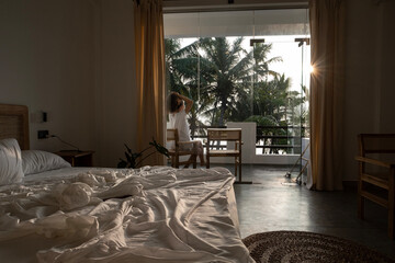 Obraz premium A young woman with long dark hair and a white T-shirt walks out onto the balcony from her luxurious bed after waking up in her hotel room. Wake-up time in a hotel room in Sri Lanka on vacation.