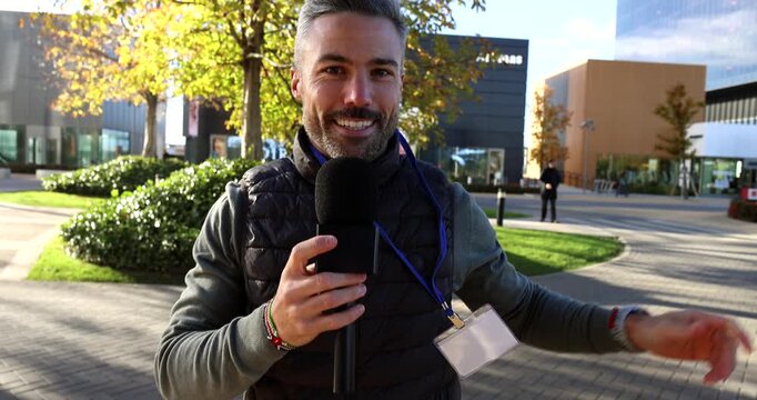 Professional male journalist with a press pass holding a microphone and speaking to the camera, reporting breaking news or presenting an event from a modern outdoor business district plaza