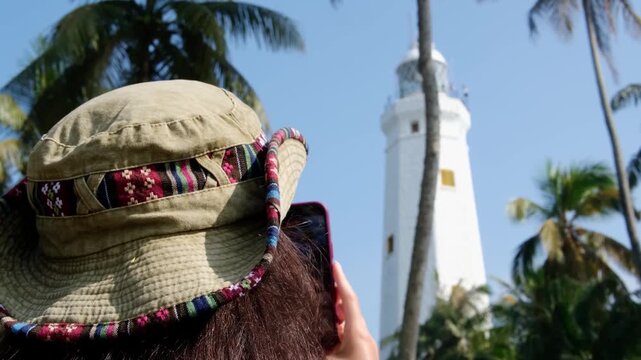 View of the white, tall lighthouse on the southern tip of the island of Sri Lanka. The lighthouse is surrounded by lush coconut trees.