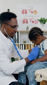 Cute 6-aged african american kid sitting on reviewing table in modern medical cabinet while good-looking friendly skillful black man-pediatrician listening her lungs breath