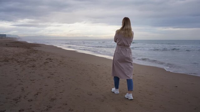 Lonely woman in trench coat walk along sandy beach and look at cold winter sea while embracing herself in cloudy and windy weather.Concept of loneliness, isolation, personal space and inner world