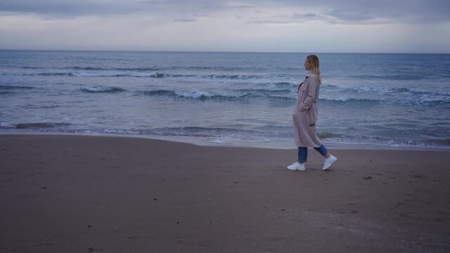 Melancholic woman in a trench coat walking alone on an empty beach in autumn.Concept of loneliness, isolation, personal space and inner world, emotional storytelling and mental health topics.