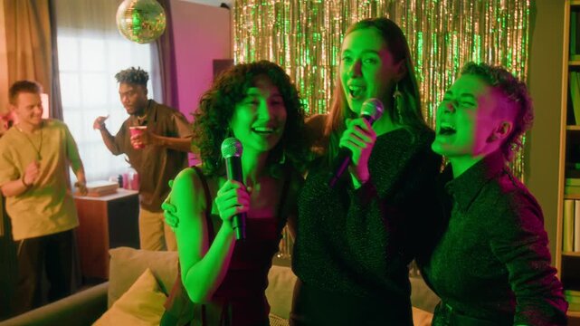 Zoom in view of three joyful gen Z female friends singing karaoke at party in home setting, two men dancing in background