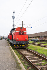 Obraz premium a vintage red diesel locomotive with yellow hazard stripes stationed on railway tracks next to an old station building under a grey sky.