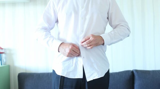 A man buttons his crisp white shirt while standing in a softly lit room, conveying quiet focus, personal care, and the ritual of daily readiness.
